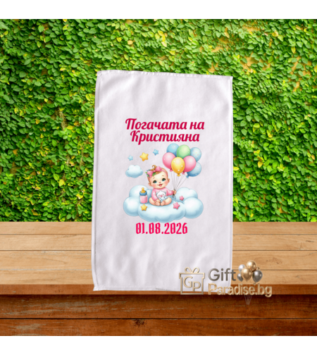 Малка бяла хавлиена кърпа 30х50 см за бебешка погача, снимана върху дървен плот пред зелена стена, с розов печат Погачата на Кристияна, дата и илюстрация на бебе с цветни балони на облак.